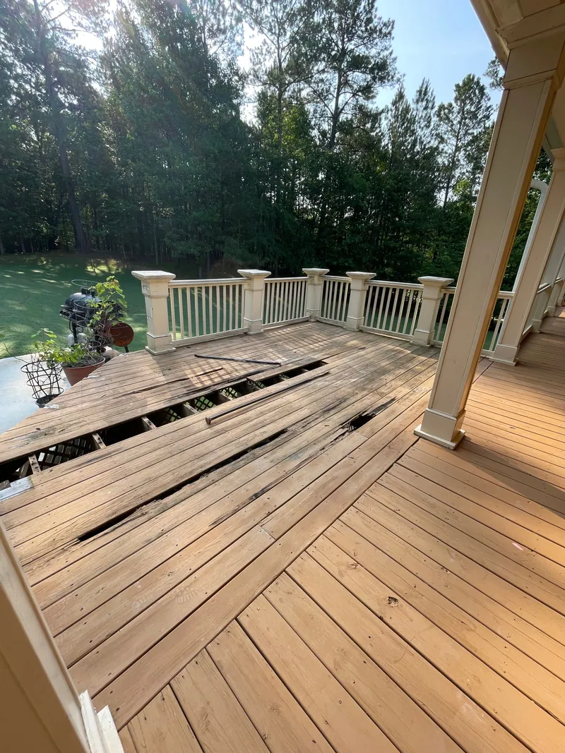 Wood deck with a run of floorboards pried up, exposing the joists underneath. Painted rail and columns behind it and pine woods at the edge of the yard.