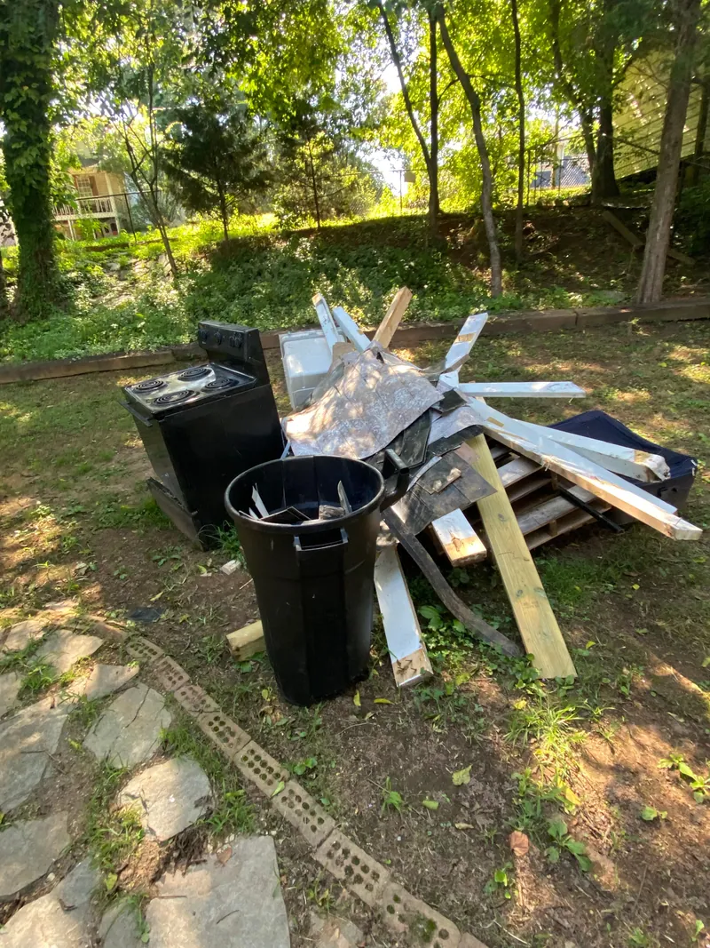 Junk piled on a shaded lawn: a black electric range, a plastic trash can, a wood pallet, cut lumber and stacks of white siding trim.