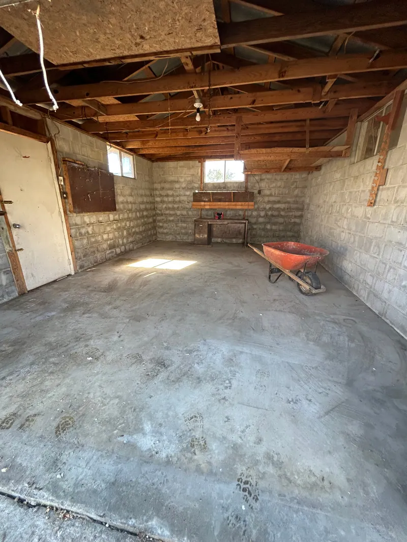 The same garage emptied to a swept concrete floor under the same rafters, with only the workbench and an empty wheelbarrow left against the block wall.