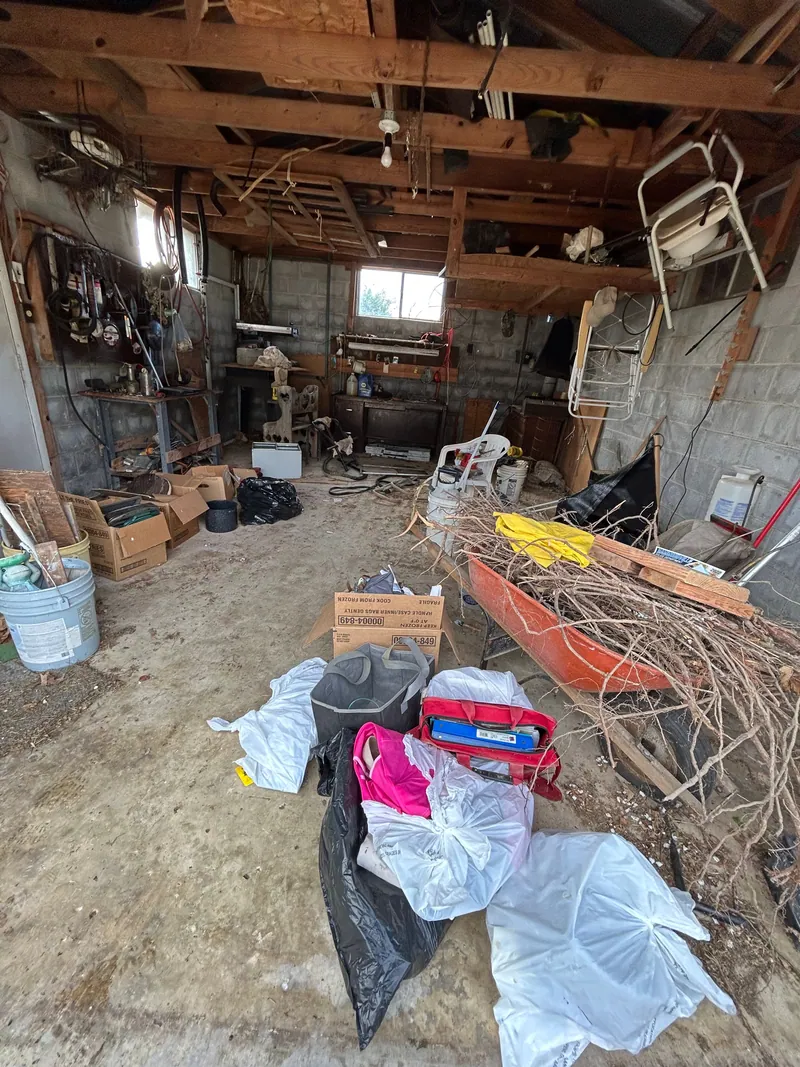 Detached garage packed with stored things: cardboard boxes, buckets, bagged items and a wheelbarrow loaded with cut brush, under open rafters and concrete block walls.