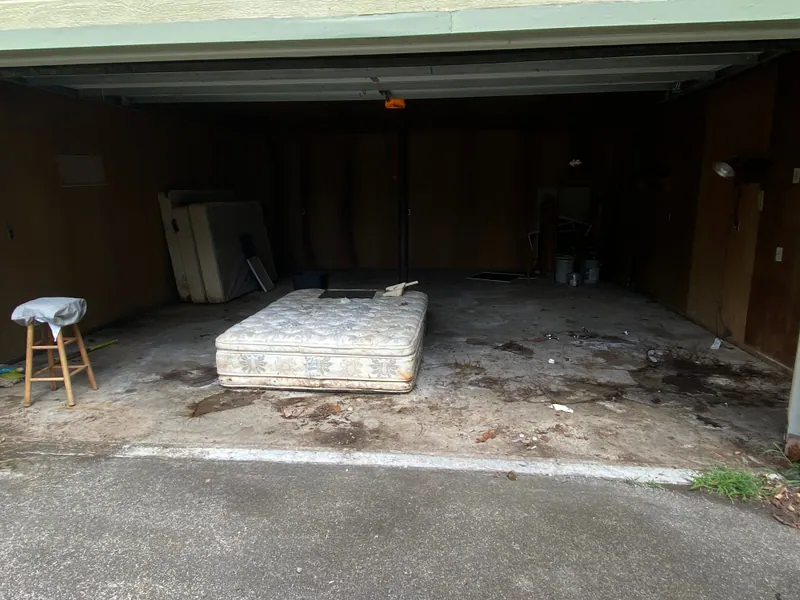 Garage with a stained mattress lying on the concrete floor, a wooden stool by the wall and panels leaning in the corner, with the door raised to the driveway.