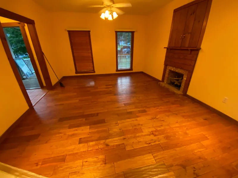 The same living room emptied to clear hardwood floor in front of the brick fireplace, with a broom left standing by the door.