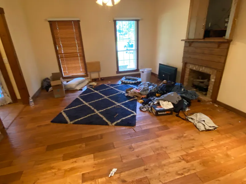 Living room with coats, clothing and a pet bed heaped on the hardwood floor beside a blue and white rug, in front of a brick fireplace.