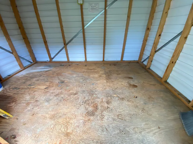 The same shed emptied to a bare plywood floor between the wall studs, with a push broom at the edge of the frame.