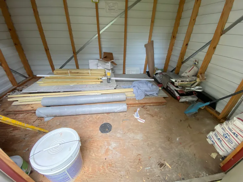 Metal shed stacked with lumber and trim, rolls of carpet, bags of mortar mix and a paint bucket across the plywood floor.