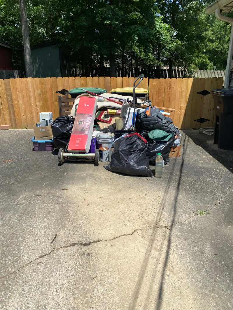 Household junk stacked on a concrete driveway against a wood privacy fence: black bags, plastic totes, buckets, rolled carpet, a hand truck and wheeled trash cans.