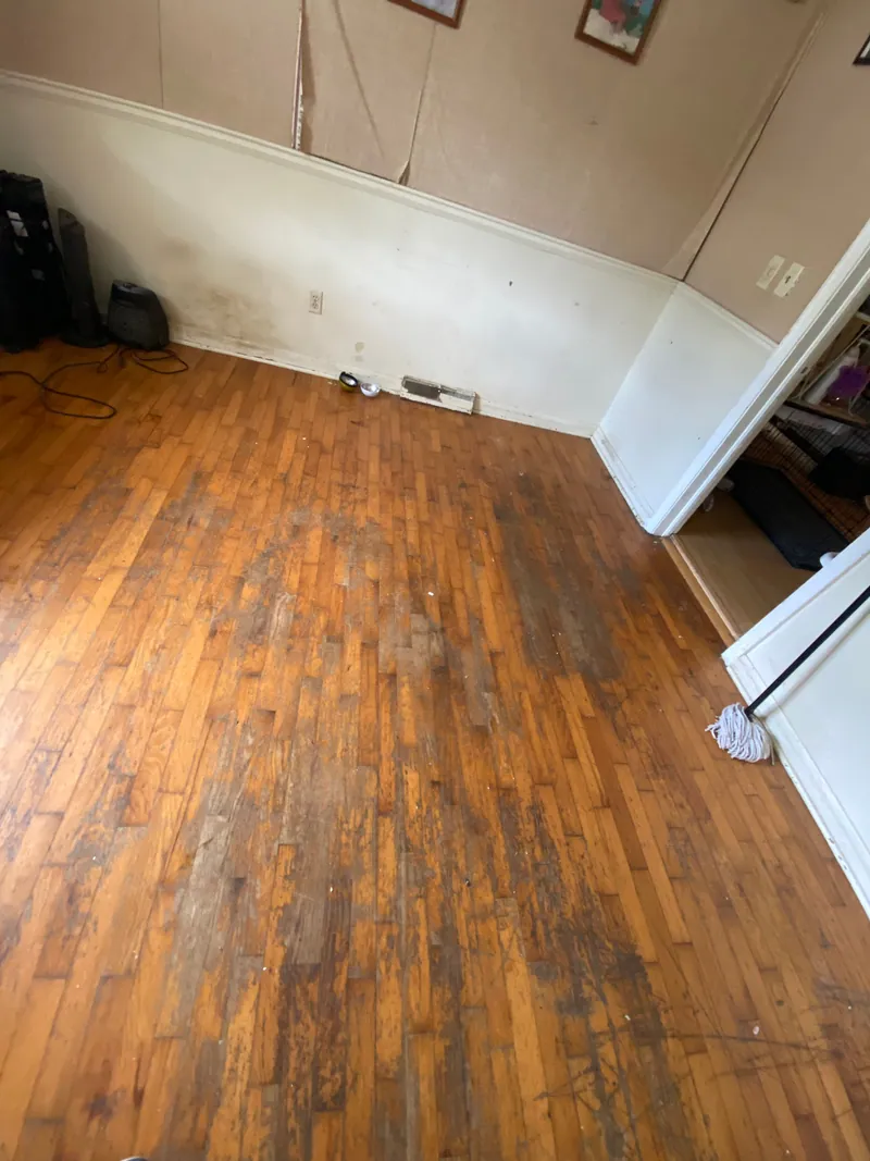 Room cleared to a bare hardwood floor, with a mop leaning by the door frame and dark marks on the boards where a pile had been stacked.