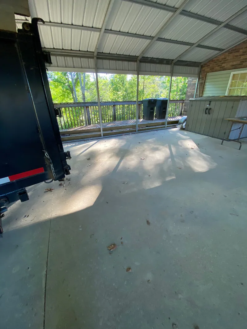 The same carport emptied to clear concrete, with the open tailgate of a black dump trailer backed in at the left.