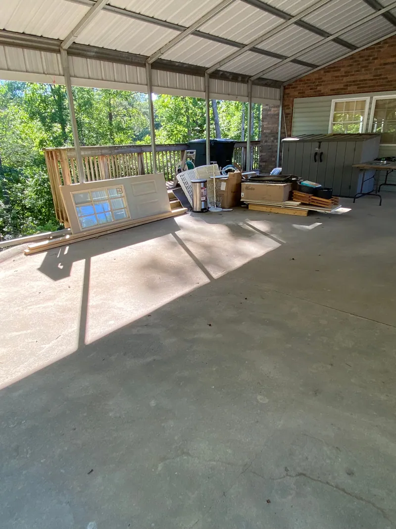 Carport stacked along the back rail with a paneled door, framing lumber, cabinet doors, cardboard boxes and a laundry basket.