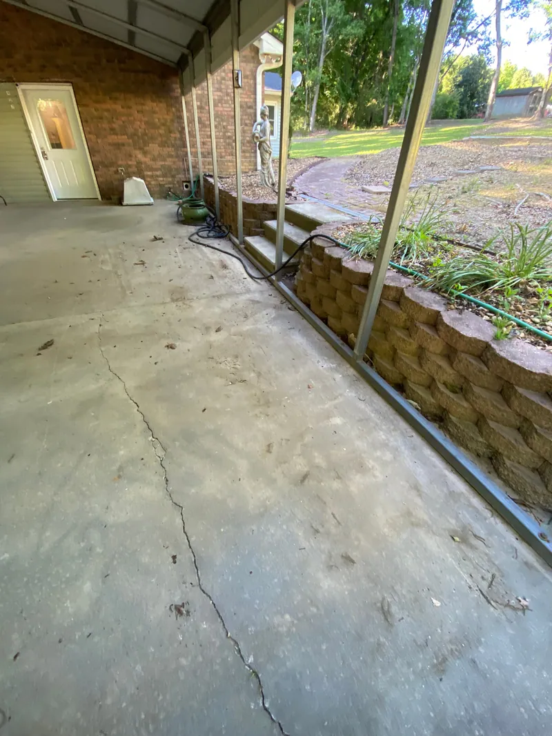 The same carport corner cleared to swept concrete, with only a garden hose and a sprayer left against the brick wall.