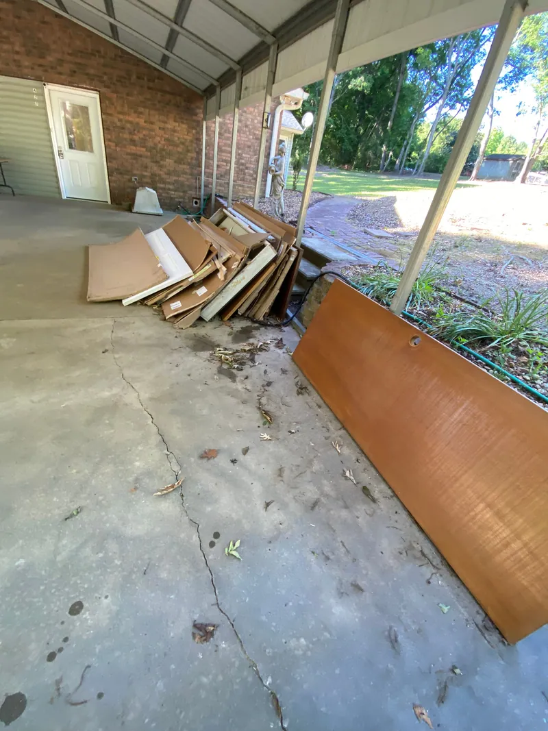 Flattened cardboard, cabinet doors and a large wood panel stacked on the concrete floor of a covered carport beside a brick wall.